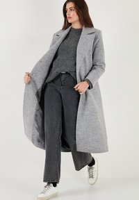 Grijze wolmix jas met een tailored ontwerp, notch-lapels en dubbelknopen, gedragen over een grijze trui en donkere spijkerbroek.