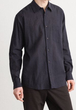 Homme portant une chemise bleu marine foncé à manches longues avec de subtiles rayures verticales et un pantalon foncé, mains relâchées sur les côtés.
