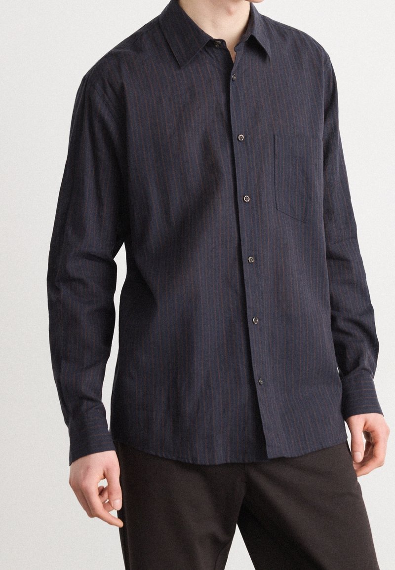 Homme portant une chemise bleu marine foncé à manches longues avec de subtiles rayures verticales et un pantalon foncé, mains relâchées sur les côtés.