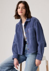Blauwe corduroy jas met een kraag, knoopsluiting, borstzak en een oversized pasvorm, gecombineerd met lichtblauwe spijkerbroeken.