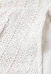 Witte kanten stof met ingewikkelde patronen, waaronder geometrische vormen en getextureerde gebieden, gelaagd op een gladde witte achtergrond.