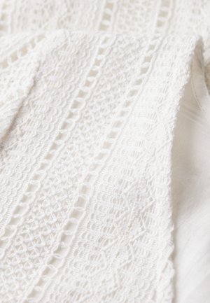 Tissu en dentelle blanche présentant des motifs complexes, comprenant des formes géométriques et des zones texturées, superposés sur un fond blanc lisse.