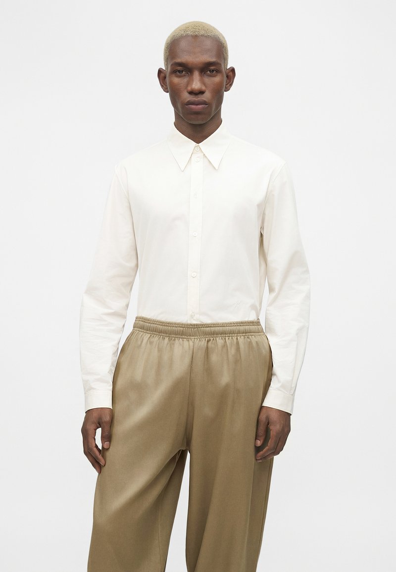 Homme aux cheveux blonds courts portant une chemise blanche boutonnée et un pantalon beige ample, debout devant un fond blanc uni.