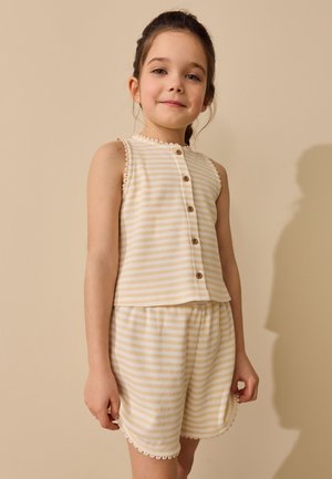 Jong meisje dat een mouwloze geel-wit gestreepte top met knopen en bijpassende korte broek draagt, staand tegen een beige achtergrond.
