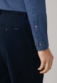 Acercamiento de una persona que lleva pantalones azul marino con un bolsillo trasero abotonado y una camisa de manga larga azul texturizada con puños abotonados.