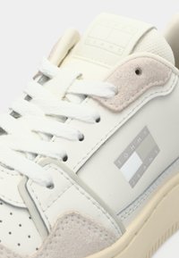 White Tommy Jeans sneaker with beige suede accents, white laces, and logo on the side and tongue, close-up view.