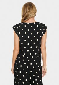 Blouse noire avec de courtes manches chauve-souris, de grands pois crème et un ourlet arrondi, fabriquée à partir d'un tissu léger et lisse.