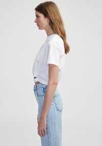 Witte cropped t-shirt met korte mouwen, geknoopt aan de zijkant; gecombineerd met lichtblauwe high-waisted jeans met zichtbare stikdetails.