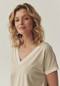 Top beige de cuello en V hecho de una tela suave, con mangas cortas y acentos a rayas en el escote y los puños. Textura lisa.