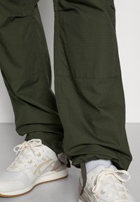 Benen in donkergroene cargo-broeken en off-white sneakers met beige en lichtbruine accenten, staand op een lichtgrijze vloer.