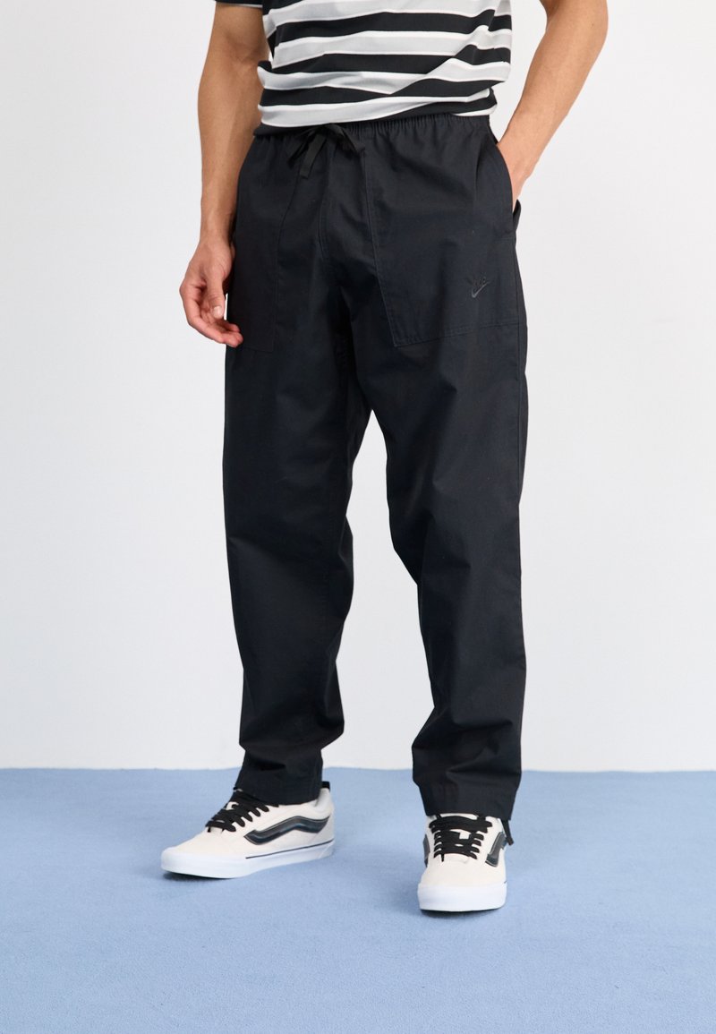 Pantaloni cargo neri con vita regolabile, tasche laterali e un sottile dettaglio del logo, abbinati a sneaker bianche con accenti neri.