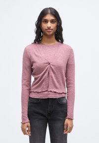 Πλεκτή μπλούζα με μακριά μανίκια σε απαλό ροζ ύφασμα με ραβδώσεις. Διαθέτει σχεδιασμό τύπου ντουμπλέ με κούμπωμα κουμπιού και βολάν στο τελείωμα. Συνδυάζεται με σκούρο τζιν.