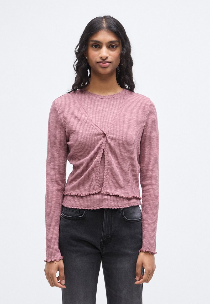 Πλεκτή μπλούζα με μακριά μανίκια σε απαλό ροζ ύφασμα με ραβδώσεις. Διαθέτει σχεδιασμό τύπου ντουμπλέ με κούμπωμα κουμπιού και βολάν στο τελείωμα. Συνδυάζεται με σκούρο τζιν.
