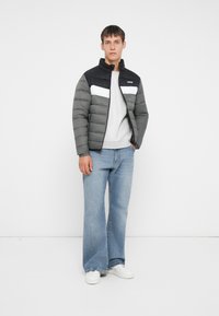 Puffervest i mørkegrå og sort med hvide detaljer, med høj krave og vandret quiltet mønster. Lysegrå sweatshirt og denim jeans.