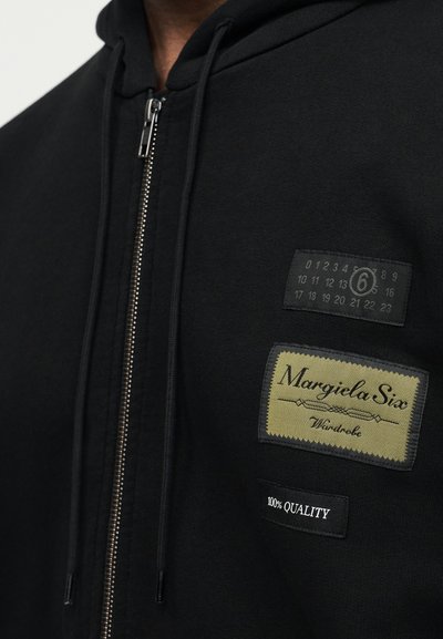 Μαύρο φούτερ με φερμουάρ, κατασκευασμένο από βαμβάκι, με δύο υφασμάτινα σχέδια: μια πράσινη ετικέτα με την επιγραφή «Margiela Six» και μια μαύρη ετικέτα που αναγράφει «100% ΠΟΙΟΤΗΤΑ».