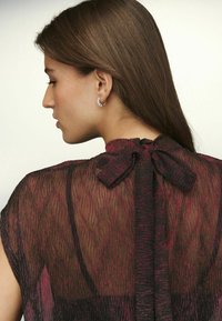 Black and pink sheer blouse with textured fabric, bow detail at the back neck, and short sleeves. Silver hoop earring visible on model.