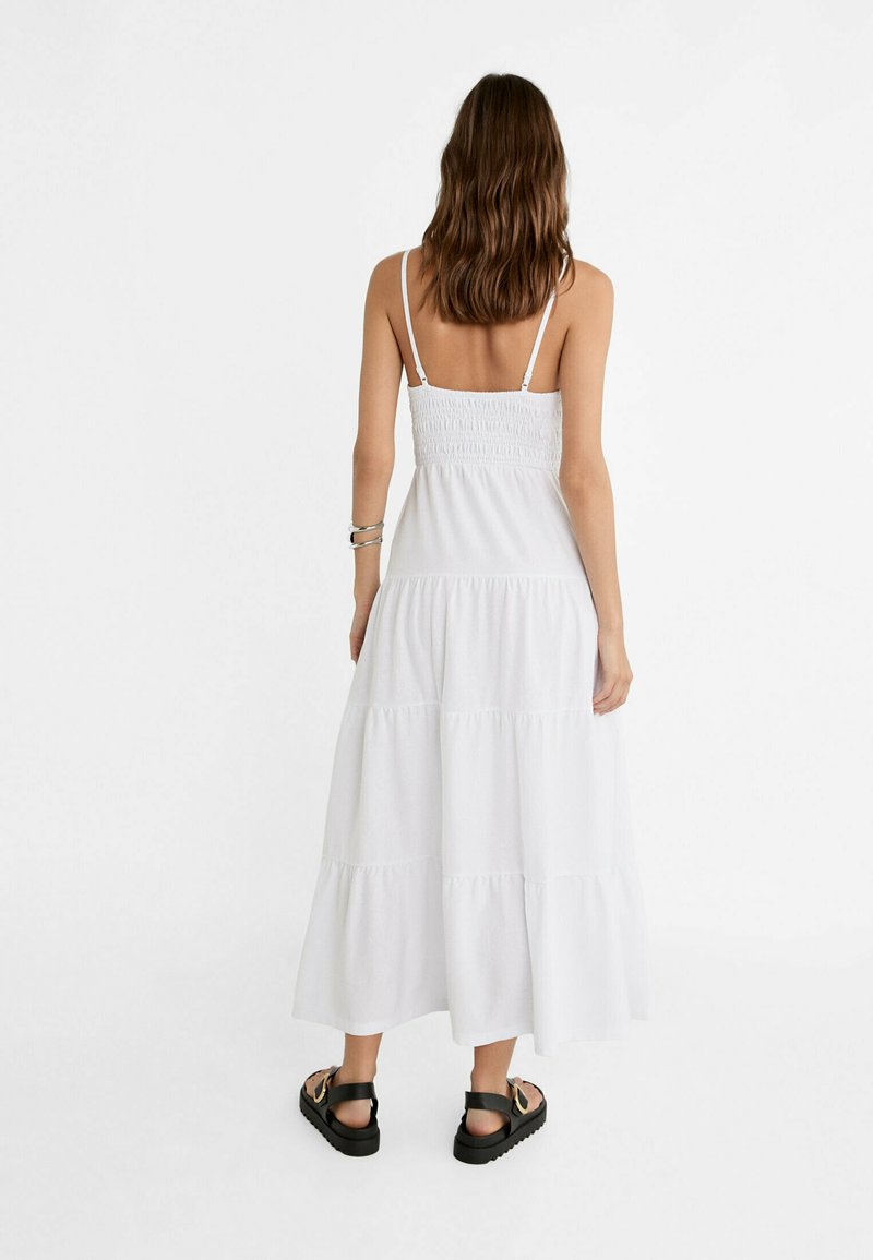Weißes ärmelloses Maxikleid mit einem gerafften Oberteil und schichteter Rockpartie. Spaghetti-Träger, leichtes Material und bequeme Passform. Schwarze Sandalen sichtbar.