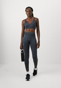 Tumši pelēks sporta krūšturis ar V-veida kakla dizainu, apvienots ar augsto jostasvietu legingiem tajā pašā krāsā, turēts ar melnu ūdens pudeli.