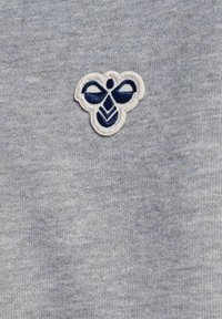 Szary materiał z małym, haftowanym logo pszczoły w granacie z białymi akcentami, umieszczonym na teksturowanym tle.