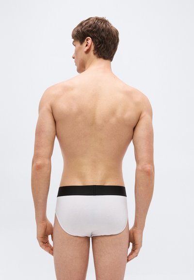Modèle masculin vu de dos portant un slip blanc avec une ceinture noire sur un fond gris clair uni.