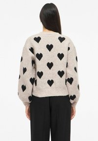 Žena s dlouhými tmavými vlasy, oblečená v béžovém svetru s černými vzory srdcí a volných černých kalhotách, zobrazená zezadu na bílém pozadí.