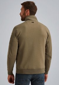 Olijfgroene, gestructureerde jas met hoge kraag en lange mouwen. Heeft een horizontaal stiksels patroon en logo-detail op de rug.