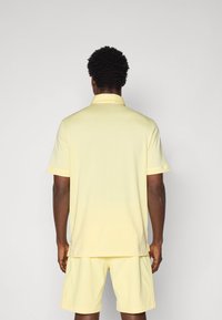 Cămașă polo galbenă cu mâneci scurte și guler, acompaniată de pantaloni scurți asortați. Textură netedă, croială lejeră, design minimalist.