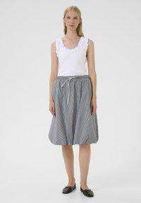 Top sin mangas blanco con cuello con volantes, combinado con una falda gris y blanca a rayas que llega a la rodilla. Unos zapatos planos negros completan el conjunto.