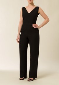 Svart ärmlös jumpsuit med V-ringning, skräddarsydd passform, vida ben och minimalistisk design, uppvisad på en modell mot en neutral bakgrund.