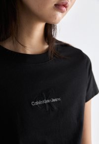 Juodas medvilnės marškinėliai su apvalia iškirpte ir trumpomis rankovėmis, su iškilniu "Calvin Klein Jeans" logotipu ant krūtinės.