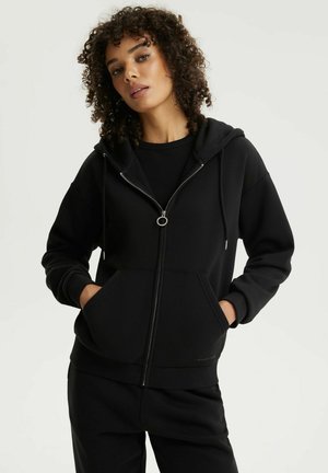 Młoda kobieta z kręconymi włosami ubrana w czarną bluzę z zamkiem i czarne spodnie, stojąca z rękami w kieszeniach na białym tle.