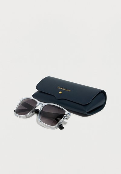 Γυαλιά ηλίου με διάφανη σκελετό και σκοτεινούς φακούς δίπλα σε μαύρη σκληρή θήκη Polo Ralph Lauren με χρυσό κουμπί και λογότυπο.