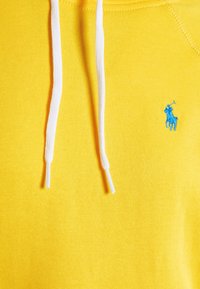 Gul hoodie i bomull med en mjuk textur, med vita dragsko och en liten broderad blå logotyp på bröstet.