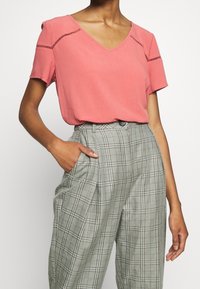 Top rosa a maniche corte con scollatura a V e dettagli ritagliati, abbinato a pantaloni a quadri grigi a vita alta con vestibilità comoda e design con pieghe.