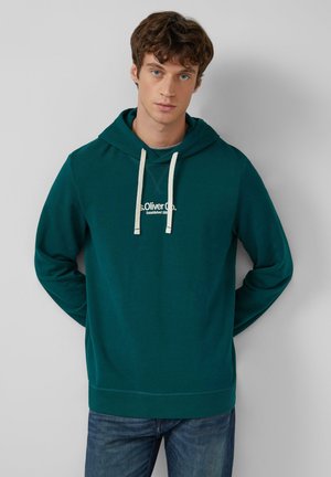 Jonge man met lichte huid en krullend bruin haar, gekleed in een donkerblauwe hoodie met witte trekkoorden en een blauwe spijkerbroek, staande tegen een grijze muur.