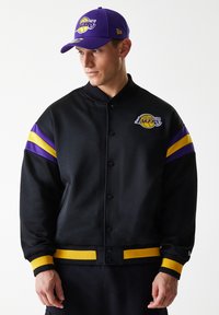 Black bomber jacket with purple and yellow stripes on sleeves, featuring a Los Angeles Lakers logo. Ribbed cuffs and waistband.