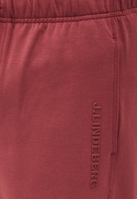 Κοντινό πλάνο σκούρων κόκκινων παντελονιών από ύφασμα με ελαστική μέση και κάθετο ανάγλυφο κείμενο "J.LINDEBERG" κατά μήκος της πλαϊνής ραφής.