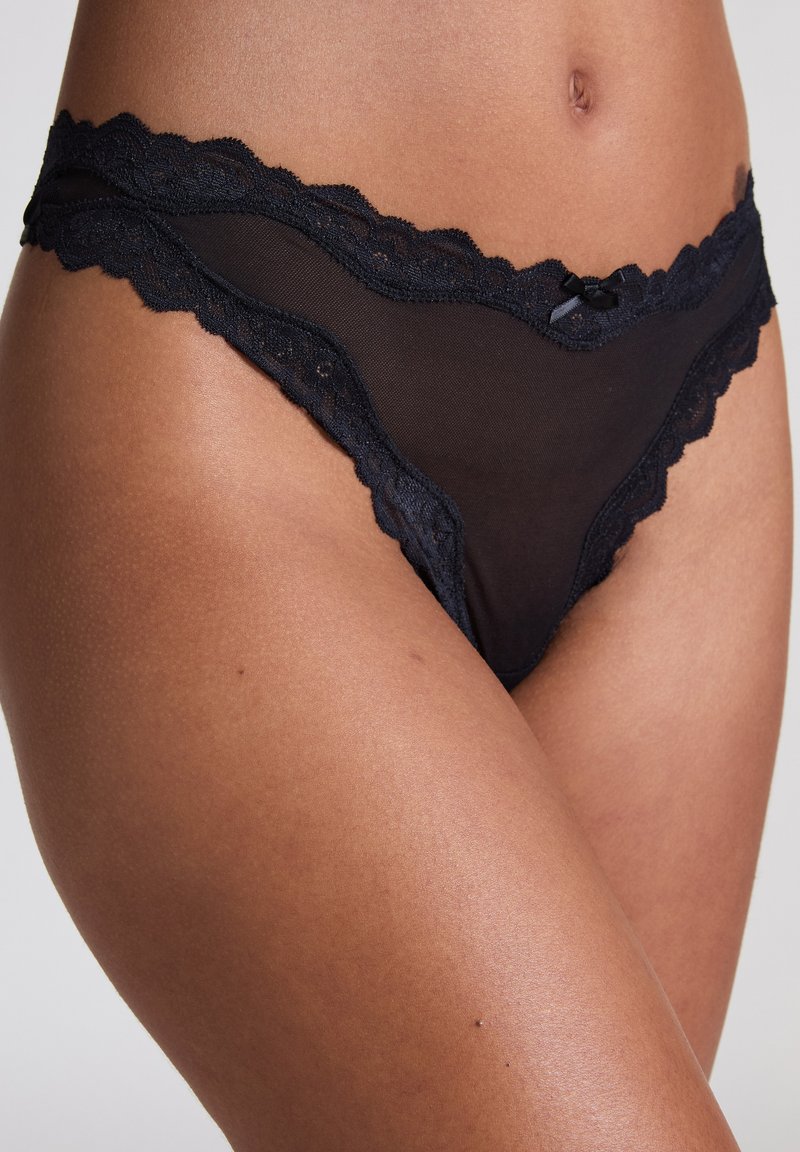 Zwarte string met kant, bestaande uit doorschijnende stof, gegolfde randen en een klein strikdetail in het midden. Kwetsbaar en licht ontwerp.