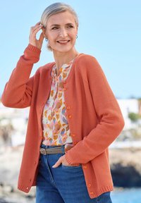 Frau, die draußen lächelt, trägt eine korallenfarbene Strickjacke, eine Bluse mit Blumenmuster, blaue Jeans und einen grauen Gürtel. Sie hat eine Hand im Haar und die andere in der Tasche.