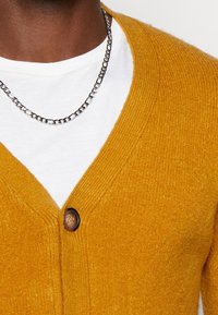 Žlutý kardigan z texturované pleteniny, s výstřihem do V a dřevěným knoflíkem vpředu, nošený přes bílé tričko.