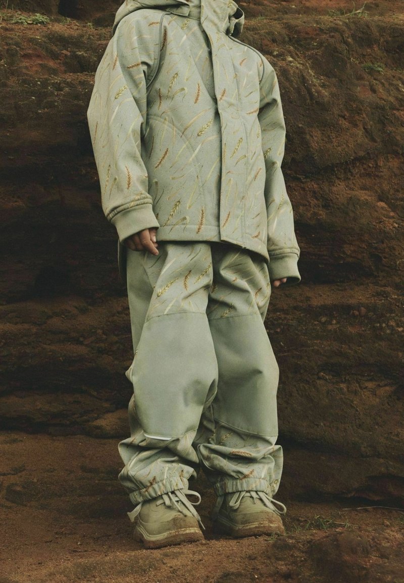 Enfant vêtue d'une veste à capuche verte et d'un pantalon assorti avec des motifs de blé, debout sur un sol terreux devant un fond rocheux.