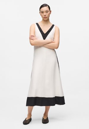 Femeie stând cu brațele încrucișate, purtând o rochie midi albă fără mâneci, cu tiv negru în formă de V la decolteu, tiv negru și pantofi slip-on împletiți negri.