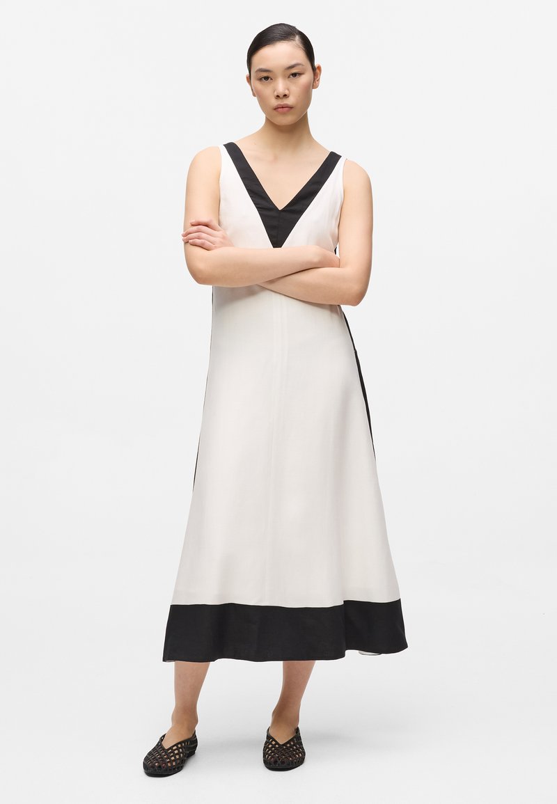 Femme debout, les bras croisés, portant une robe midi blanche sans manches avec un col en V noir, un ourlet noir et des chaussures tissées noires à enfiler.