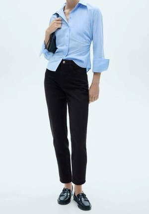 Mujer con camisa azul claro con botones, pantalones negros de talle alto, mocasines negros y lleva un pequeño bolso negro sobre un hombro.