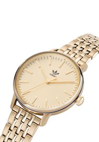 Montre dorée avec un boîtier rond, un cadran minimaliste et des index horaires fins. Elle possède un bracelet en métal tissé et une couronne sur le côté.