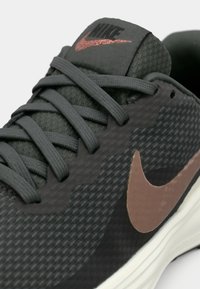 Gros plan d'une chaussure de course Nike gris foncé avec lacets noirs, dessus texturé, logo Nike métallique rose sur la languette et le côté, et semelle blanche.