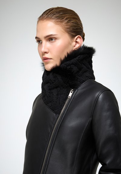 Giovane donna con capelli all'indietro, che indossa una giacca di pelle nera e una sciarpa nera lavorata a maglia alta con rifiniture in pelliccia.