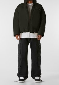 Giubbotto puffer nero con colletto alto e cerniera, indossato sopra un maglione grigio e una camicia bianca, abbinato a pantaloni cargo neri e sneakers.