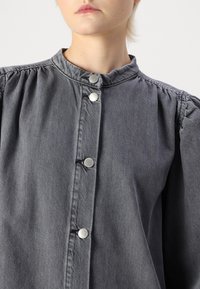 Grå denimjakke med høy krage, puffede ermer og sølvfargede trykknapper foran. Teksturert stoff med en myk overflate.