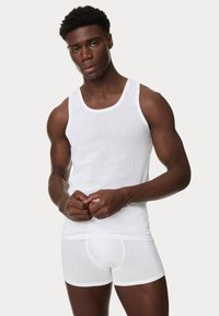 Weißes geripptes Tanktop und enge weiße Boxer-Briefs an einer Person mit neutralem Ausdruck, die glattes Material und körperbetonten Sitz zeigt.